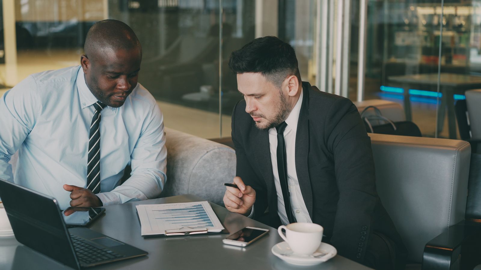 Two businessmen at a cafe table reviewing a printed financial chart